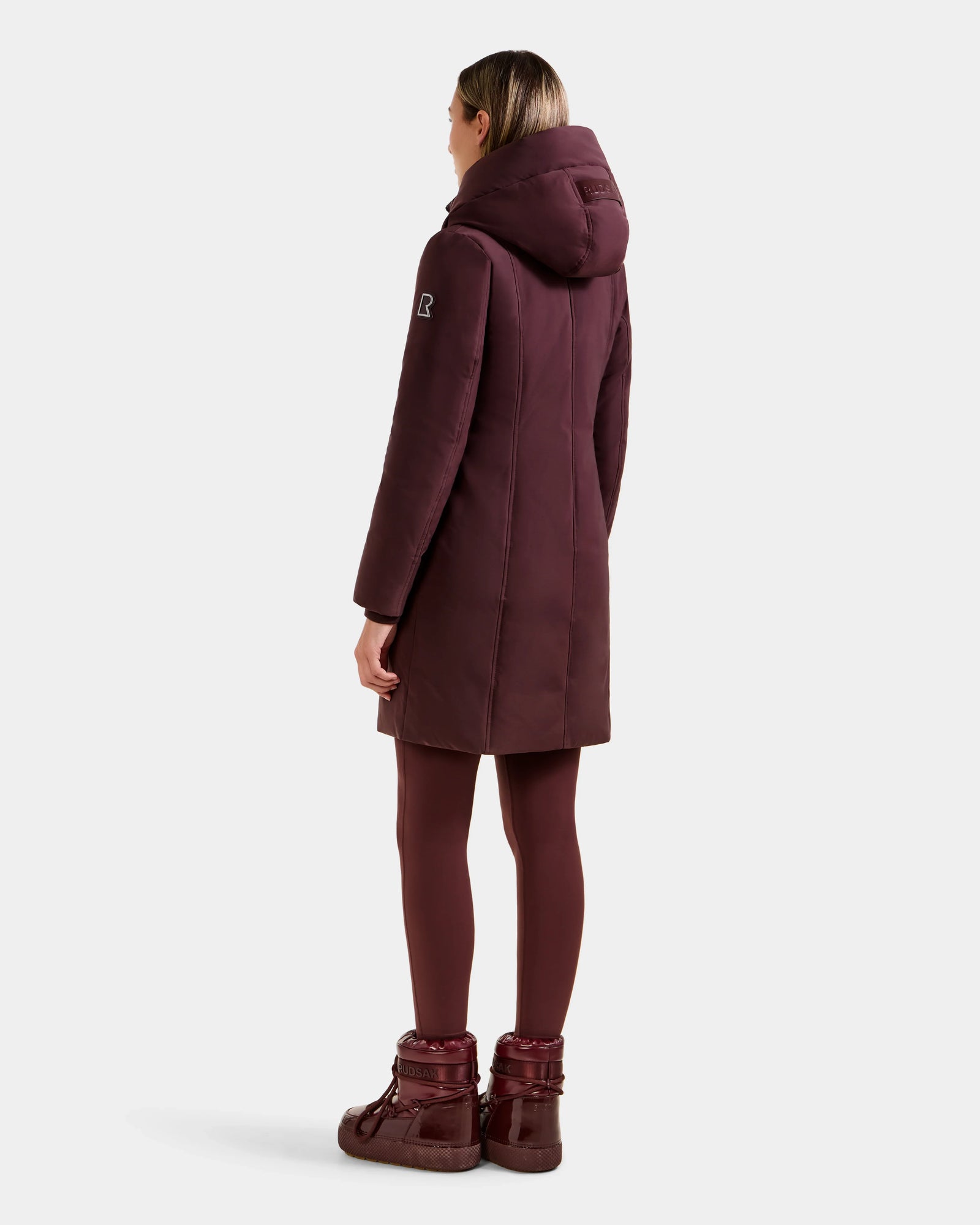 A person stands facing away, wearing the Rudsak SARINA - L125550 Bordeaux women's down parka with a hood, maroon leggings, and maroon winter boots against a plain white background.