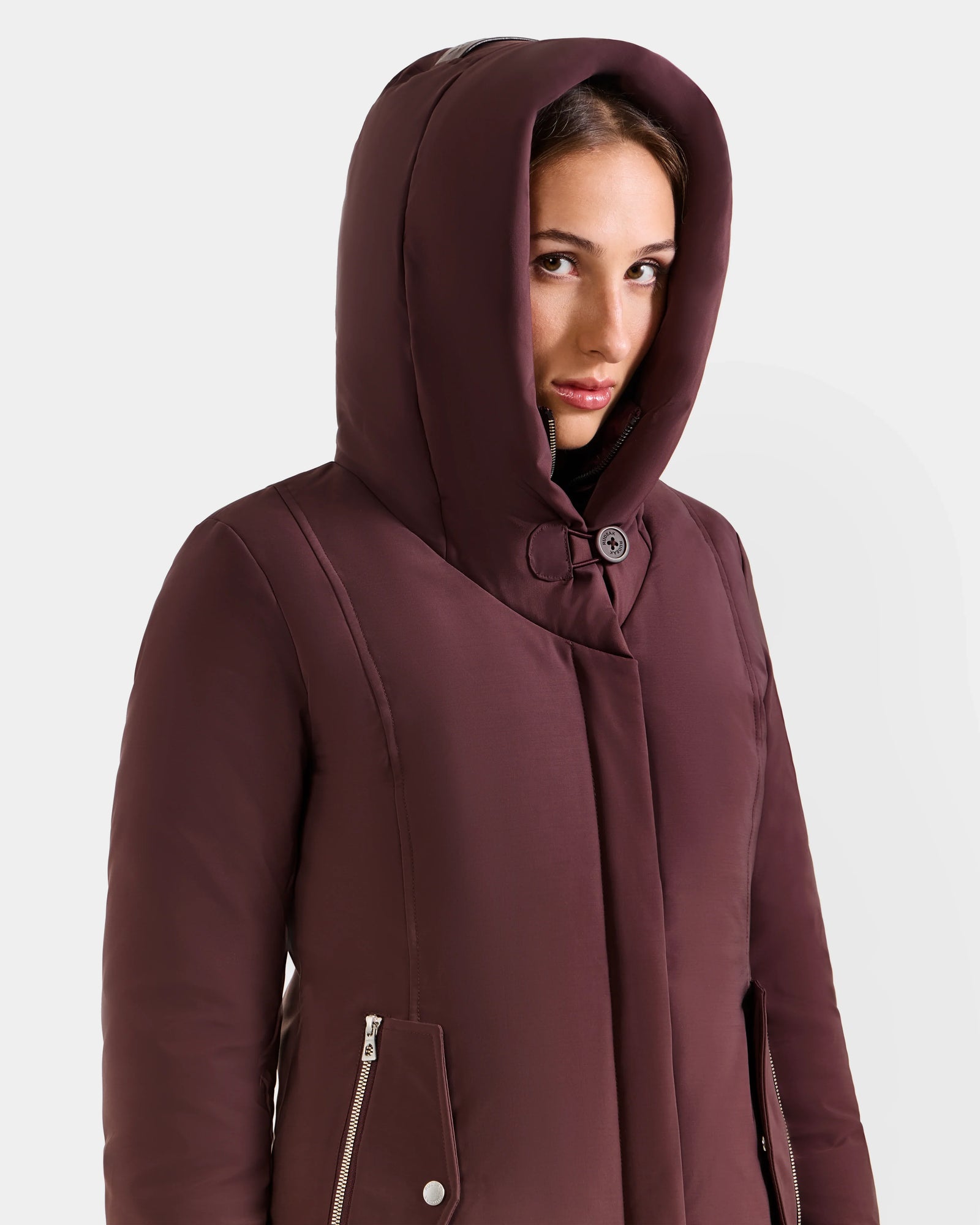 A person wearing the SARINA - L125550 Bordeaux women’s down parka by Rudsak, featuring zipped pockets, looks slightly to the side against a plain light background.
