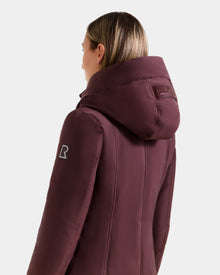 A person with light brown hair is shown from the back wearing the Rudsak SARINA - L125550 Bordeaux women's down parka, featuring a logo patch on the upper sleeve and a stitched label on the hood, against a plain white background.
