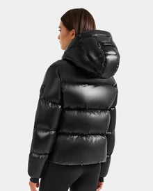 A woman with straight brown hair is seen from the back wearing the Rudsak STEFANIE S - L125585 Black women's short puffer jacket with hood, set against a plain light grey background.