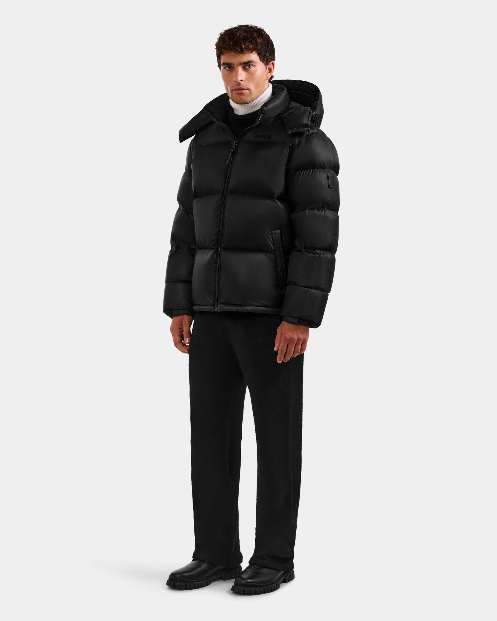 A man faces forward wearing the Rudsak STEFANO - 6125574 Black men’s down puffer jacket, paired with black pants, black shoes, and a white turtleneck against a plain light background.