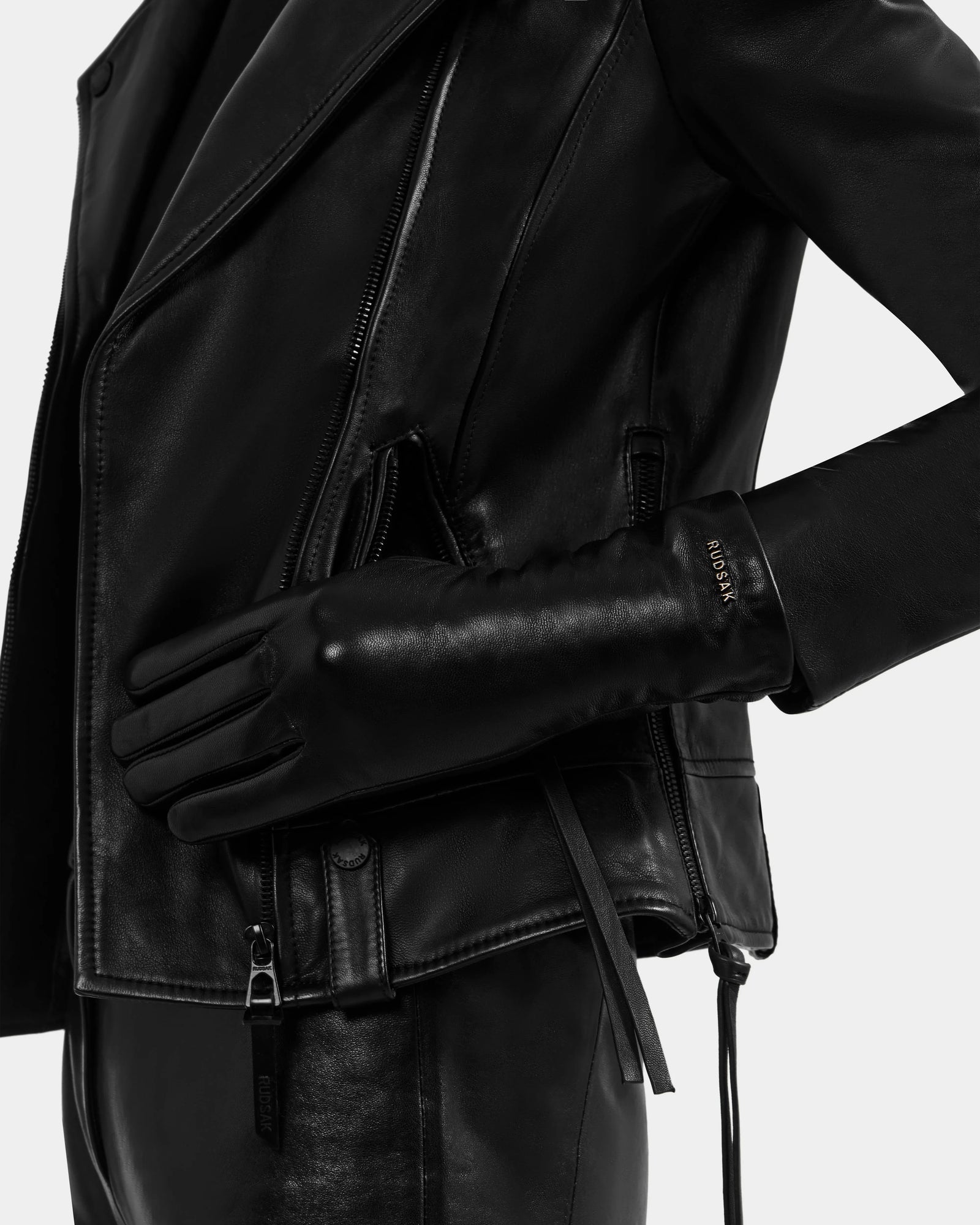 Wearing the Rudsak TORRI - L425544 Black leather jacket and women’s gloves, a person rests their gloved hand on their hip against a plain, light background, showcasing these winter essentials.