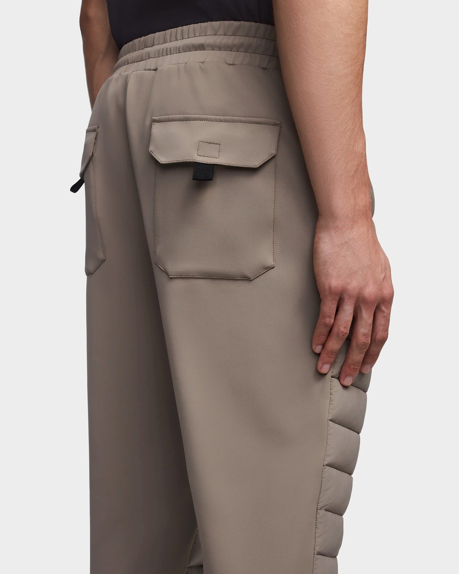 A person wearing Rudsak’s MIKE - 6523503 Cement men’s quilted pants, featuring taupe color, large flap back pockets, black pull tab, and padded side detailing. Only the lower back and one hand are visible.