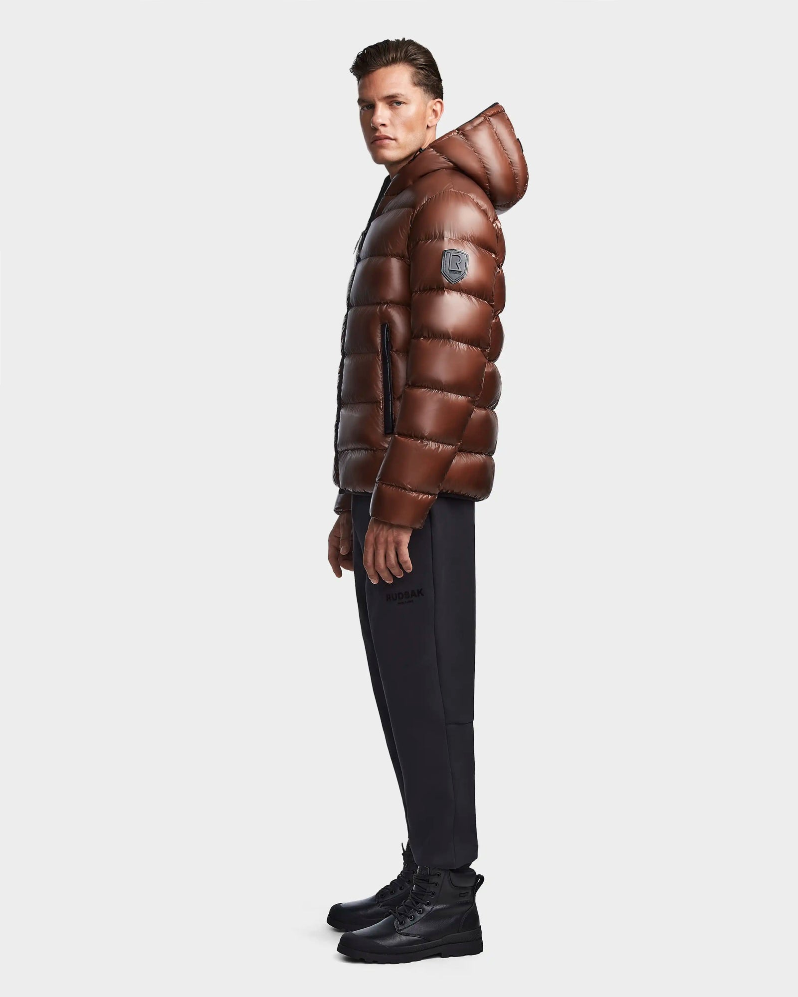 A man in profile wears the Rudsak DEMIAN S - M123508 Cohiba water and wind-resistant hooded down puffer jacket, paired with black pants and boots, set against a plain white background.