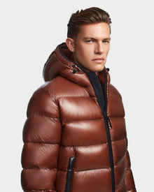 A man wearing the Rudsak DEMIAN S - M123508 Cohiba shiny brown, water and wind-resistant hooded down puffer faces slightly to the side, looking at the camera with a neutral expression against a plain background.