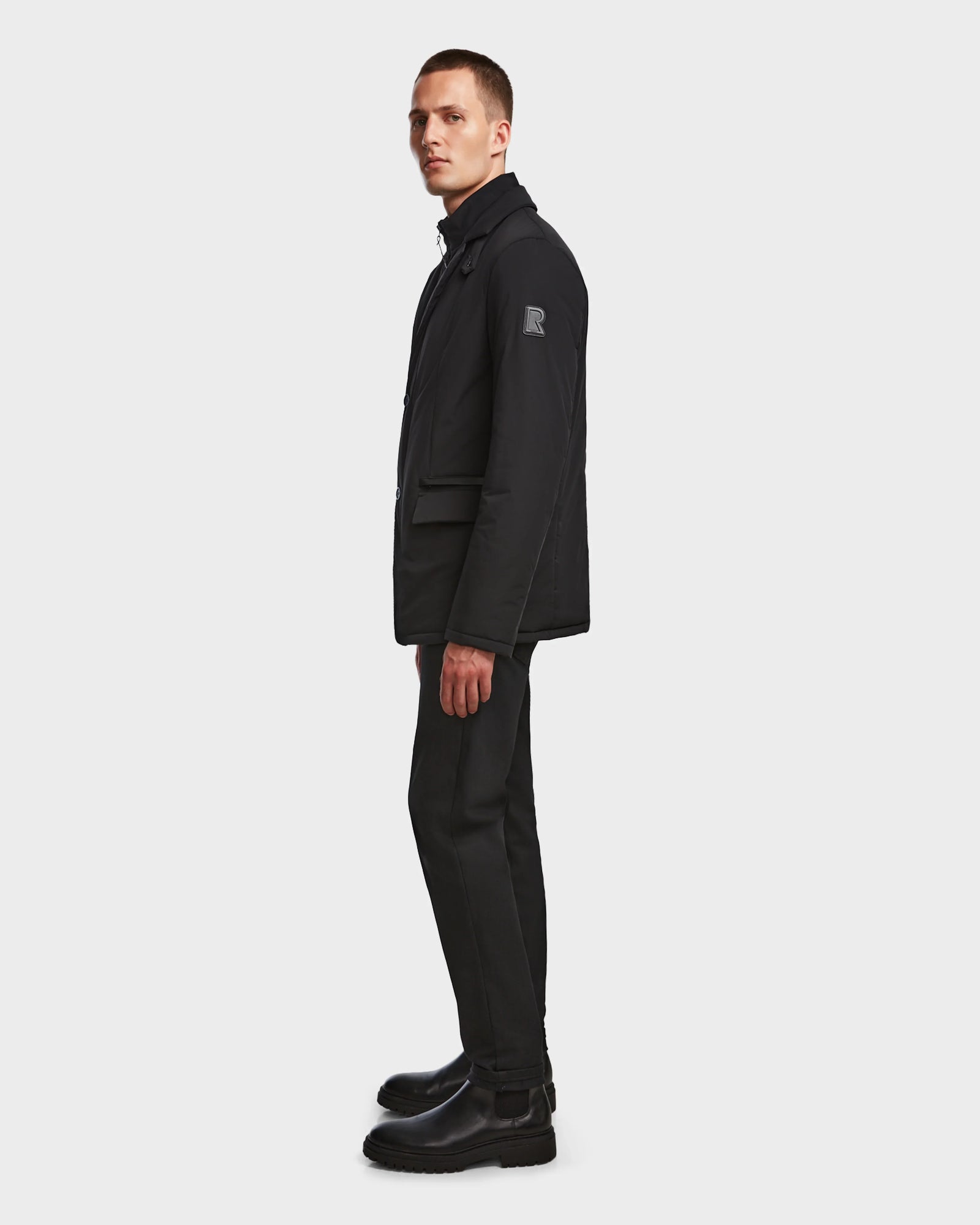 A man stands sideways against a white background, wearing the Rudsak OTTO - 6124602 Black vegan leather mens blazer-style down jacket, paired with black pants and boots. He looks forward with a neutral expression.