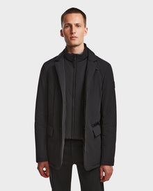 A man with short hair wears the Rudsak OTTO - 6124602 Black vegan leather mens blazer-style down jacket over a black zip-up top and black pants, standing against a plain white background.