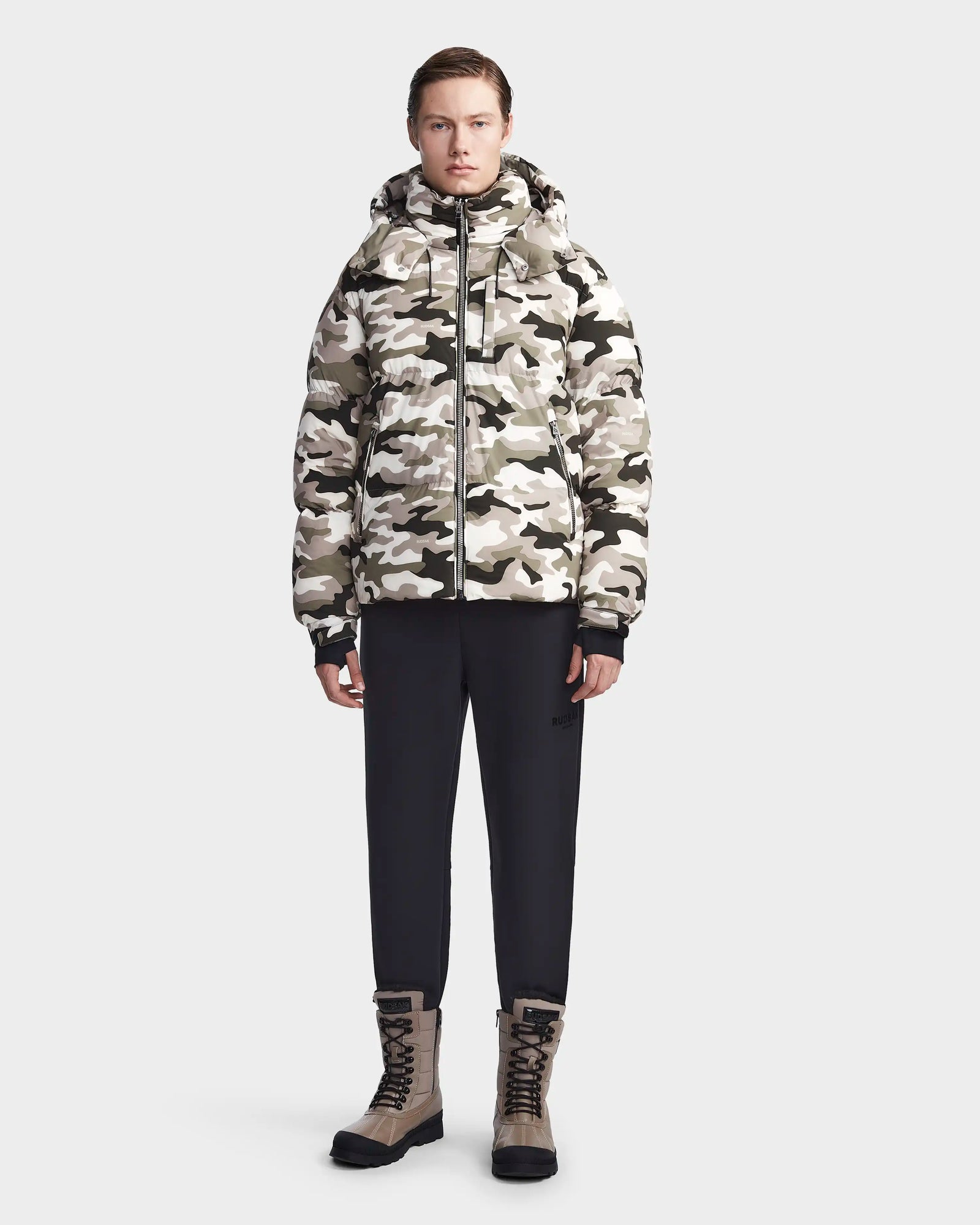 A person stands facing forward against a plain light background, wearing the Rudsak VINCENT M - 6123510 Camouflage men's down puffer in black-and-white camo, paired with black pants and tan lace-up boots.