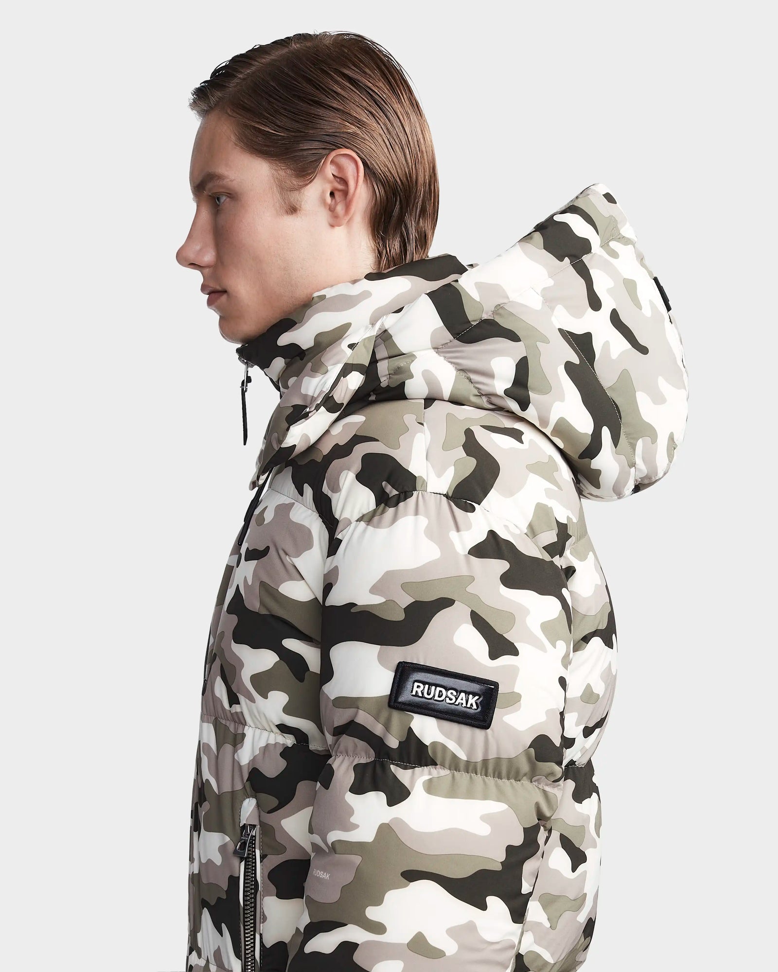 A person in profile wears the Rudsak VINCENT M - 6123510 Camouflage men’s water-repellent down puffer jacket with a visible Rudsak patch on the sleeve, standing against a plain light background.