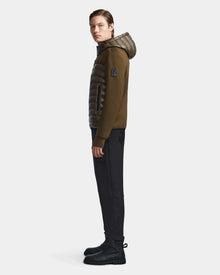 A man stands in profile against a white background wearing the Rudsak DURIN S - M123518 Light Olive mens hooded jacket with lightweight down insulation, black sleeves, waterproof zippers, black pants, and boots.