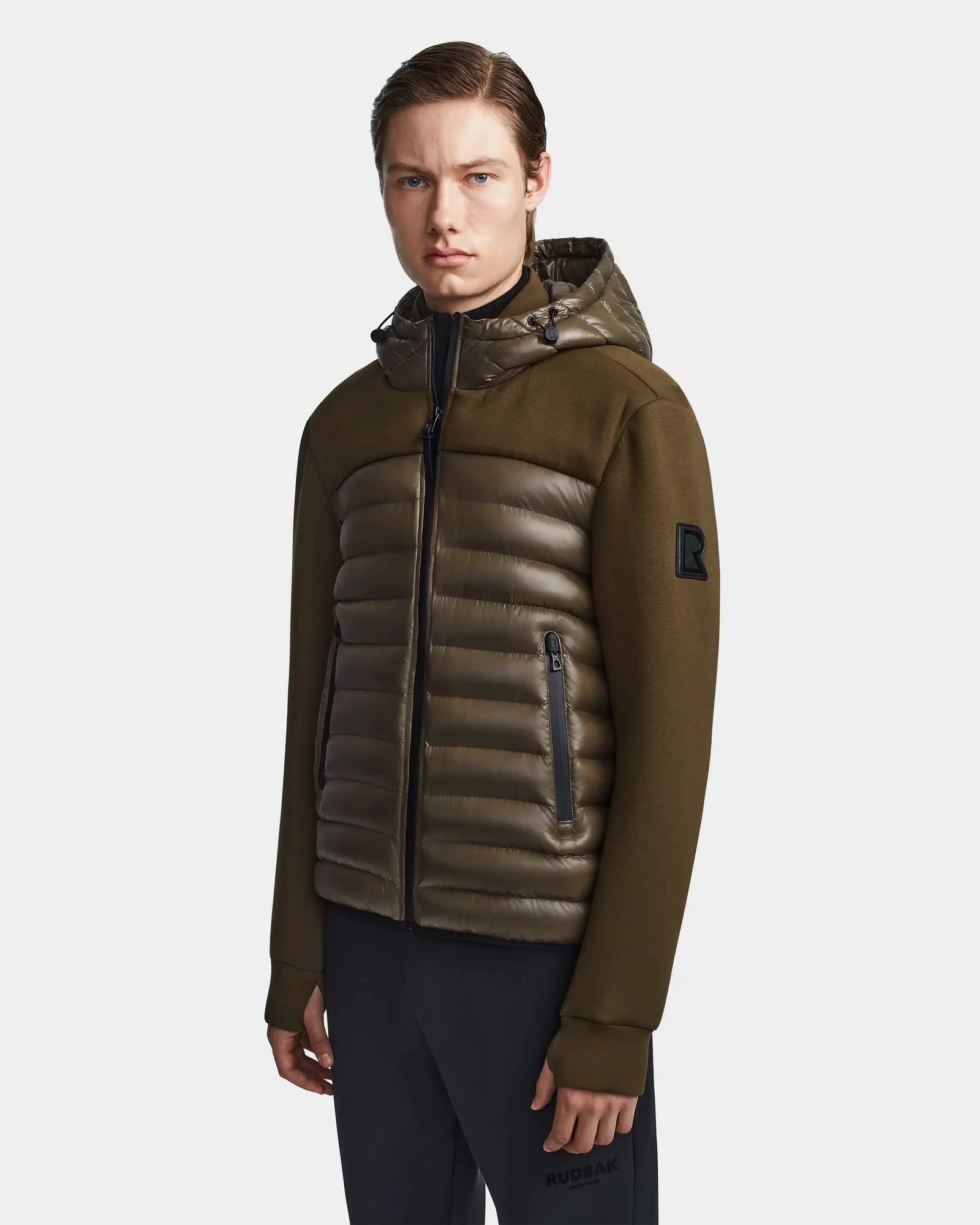 A young man with straight brown hair wears the Rudsak DURIN S - M123518 mens hooded jacket in light olive, featuring quilted panels, waterproof zippers, and lightweight down insulation, standing against a plain light background.