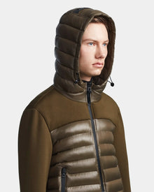 A young man wears the Rudsak DURIN S - M123518 Light Olive mens hooded jacket with horizontal quilting and a front zipper. The lightweight down jacket stands out against a plain, light background as he looks slightly to the side.