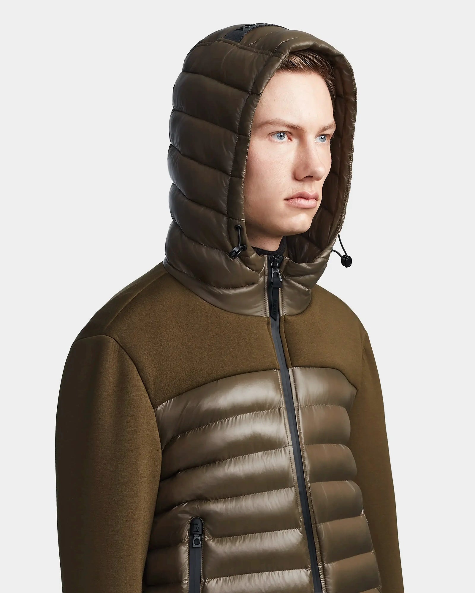 A young man wears the Rudsak DURIN S - M123518 Light Olive mens hooded jacket with horizontal quilting and a front zipper. The lightweight down jacket stands out against a plain, light background as he looks slightly to the side.