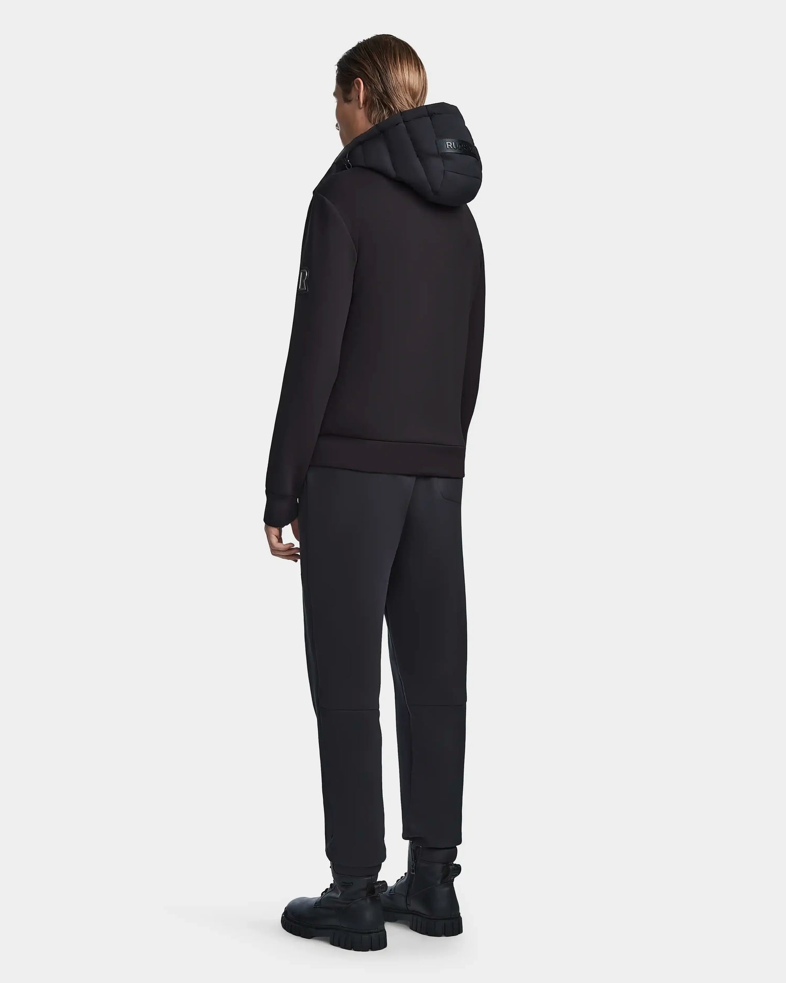 A person faces away wearing the Rudsak LAURENZO - M123514 Black neoprene jacket with removable hood, black pants, and chunky black boots, set against a plain white background.