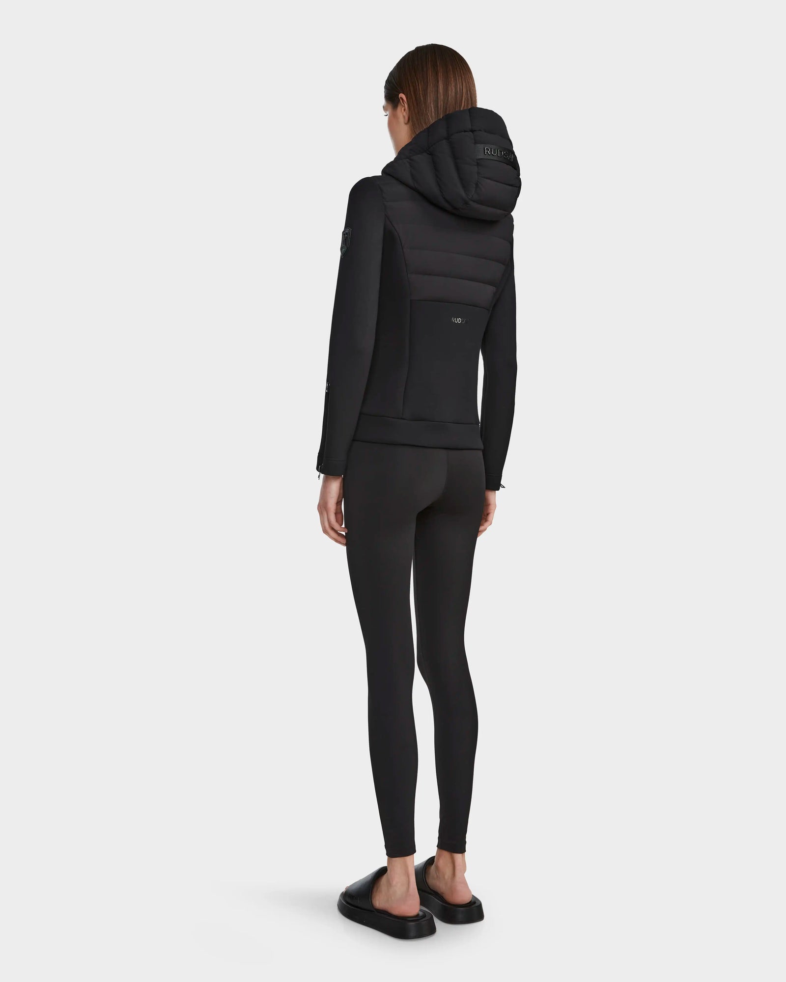 A woman faces away, wearing the RUDSAK MABS - 8122553 BLACK lightweight activewear jacket with black leggings and slide sandals against a plain white background.