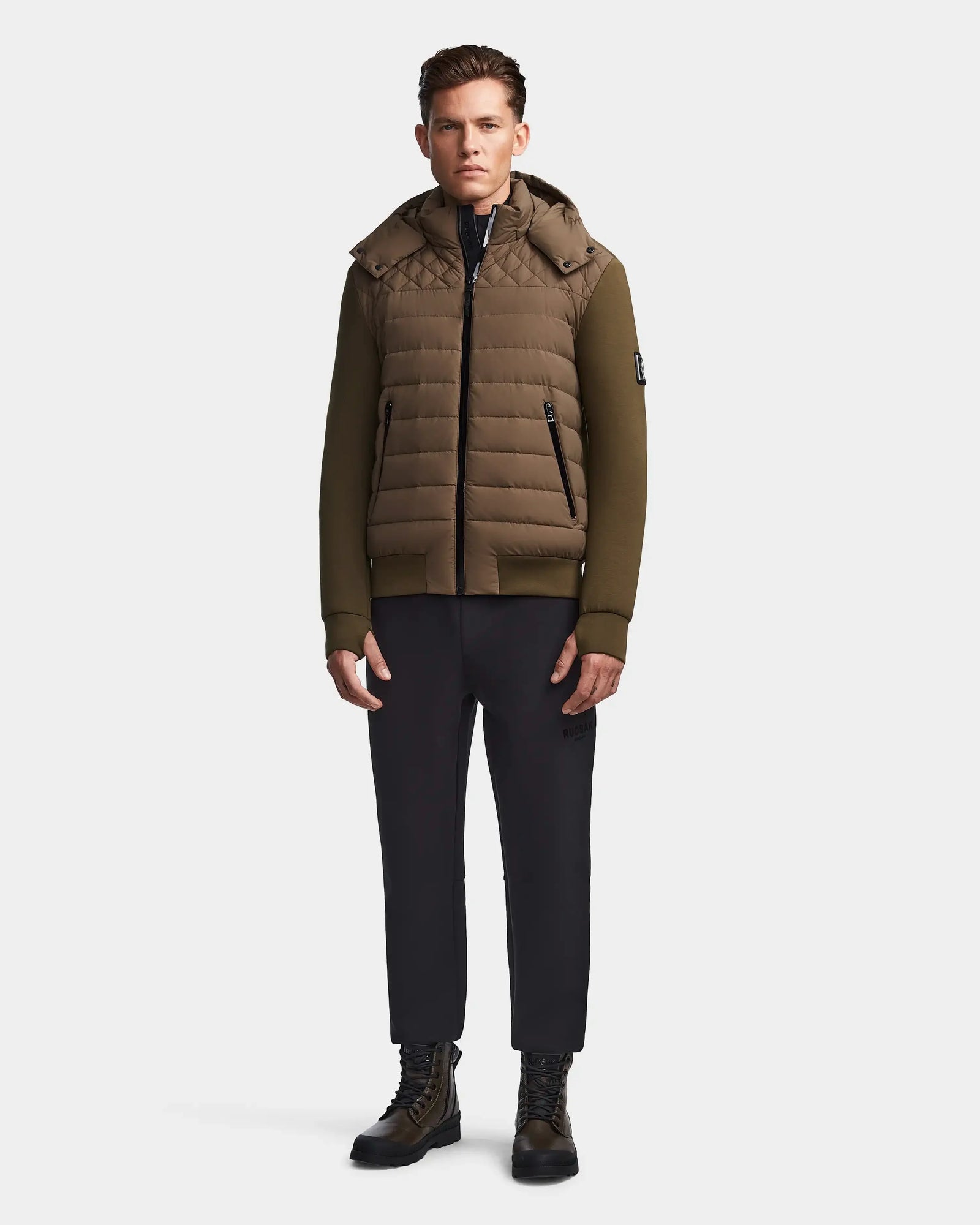 A man stands facing forward, wearing the Rudsak LAURENZO - M123514 light olive quilted hooded jacket with black pants and lace-up boots, against a plain white background.