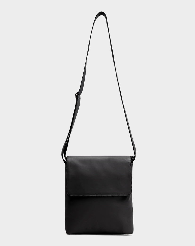 The Rudsak MADDY - 8322512 BLACK is a sleek black leather messenger bag with a long adjustable strap and front flap closure, displayed in minimalist style against a plain light gray background.