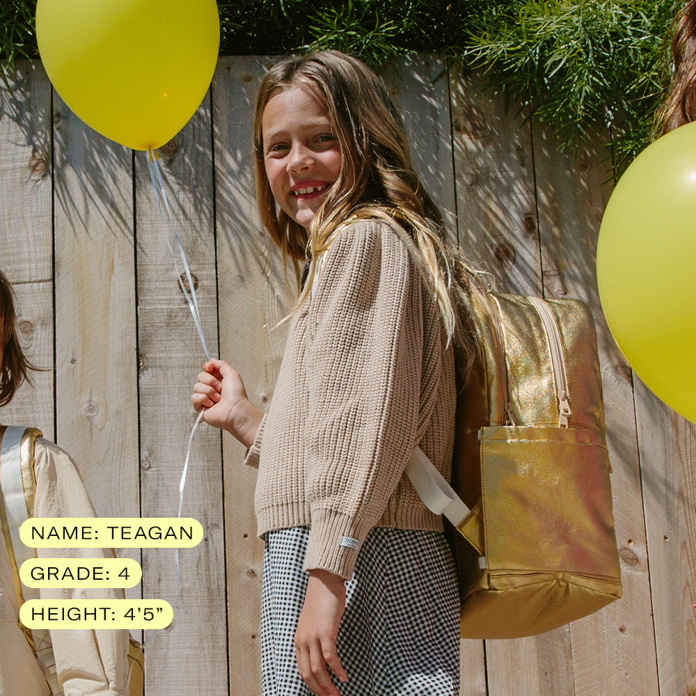 4th grade girl smiling and holding a ballon wearing state bags kane kids double pocket backpack in metallic gold 