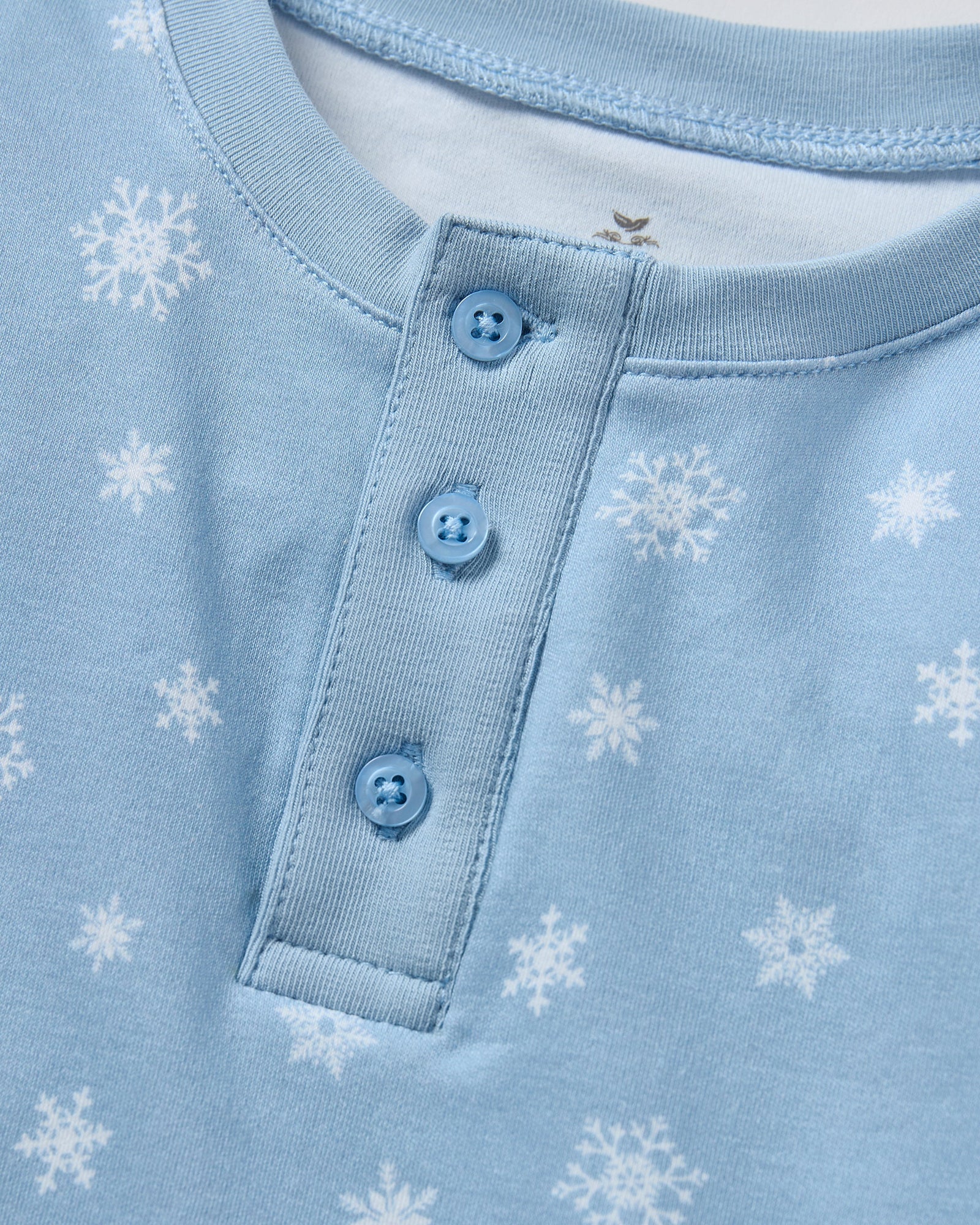 The Kid's Pima Snug Fit Pajama Set in Periwinkle Wonderland features light blue, long-sleeve pajamas with white snowflakes, buttoned neckline, matching elastic waist pants, and cuffed ankles. Shown laid flat on a white background.