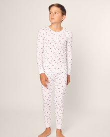A child stands barefoot against a plain background, wearing Petite Plumes Kids Pima Snug Fit Pajama Set in Apres Ski. The snug long sleeve top features a playful pattern with small trees, tents, and campfires, crafted from soft Pima cotton with a classic button-up style.