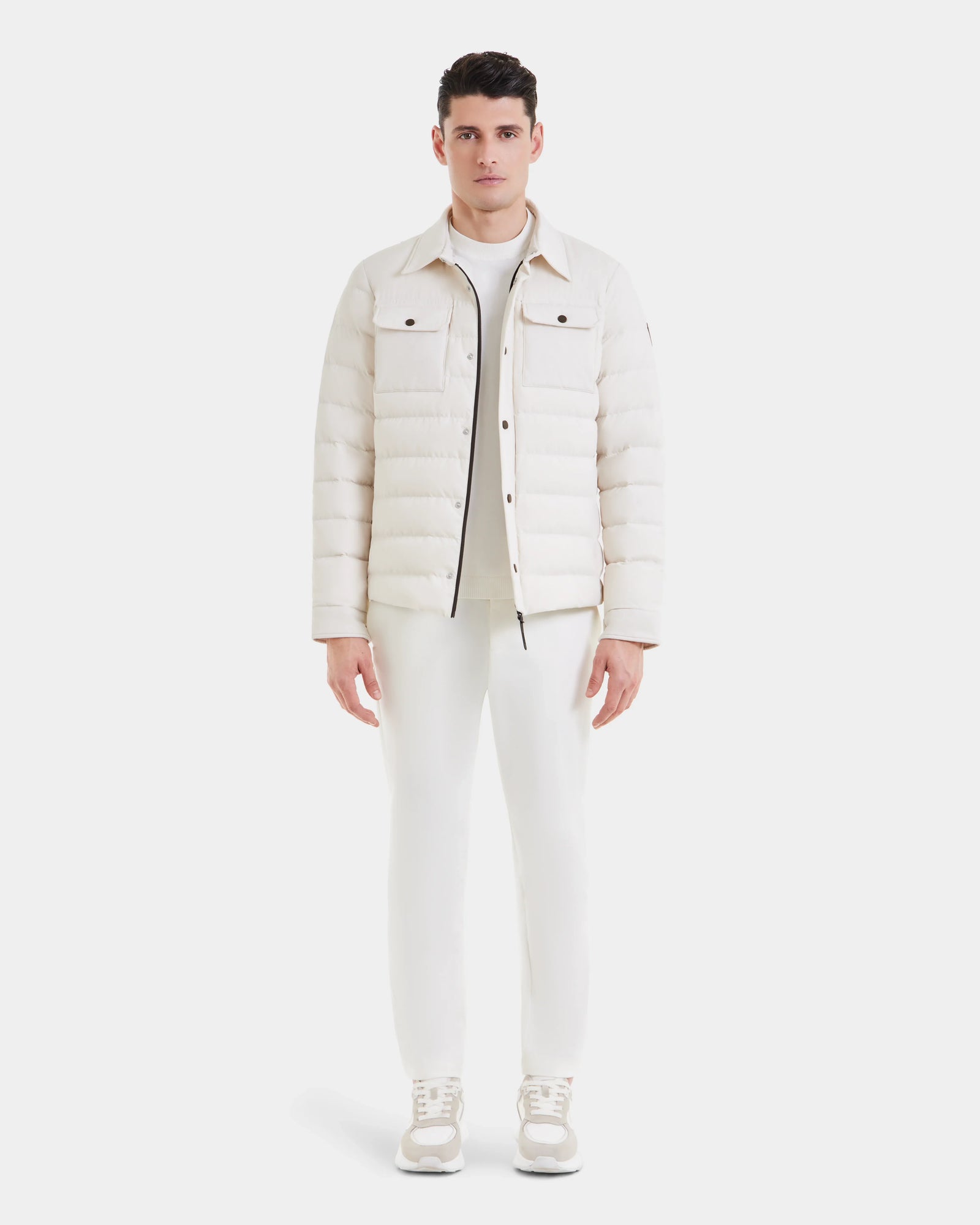 A man faces forward against a plain white background, wearing Rudsak’s AZIEL - M124521 Vanilla puffer shirt jacket, paired with white pants, a white top, and white sneakers.