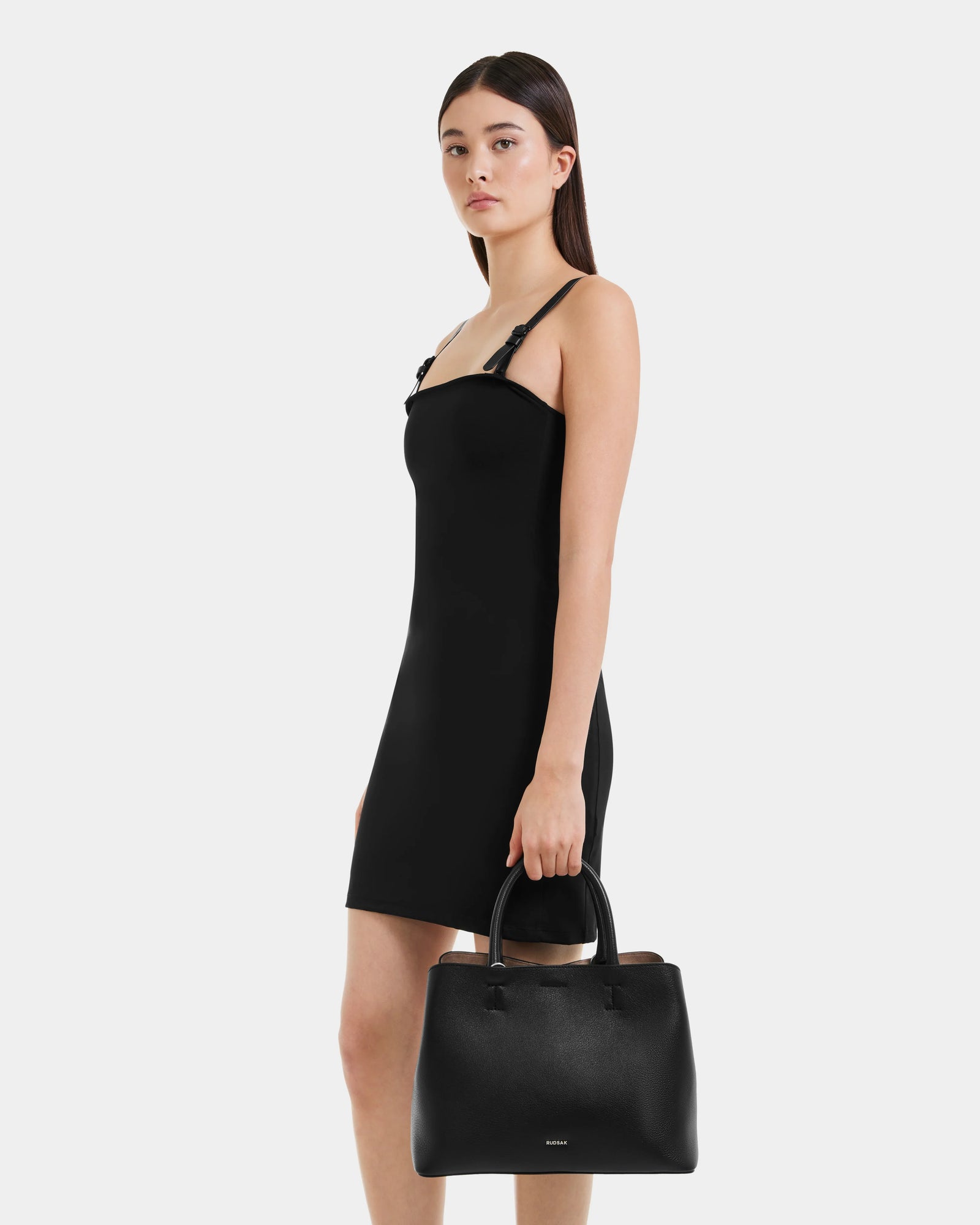 A woman in a sleeveless black dress holds the Rudsak CATIRA S - 8325006 Black leather tote by her side. She has long, straight dark hair and stands before a plain white background.