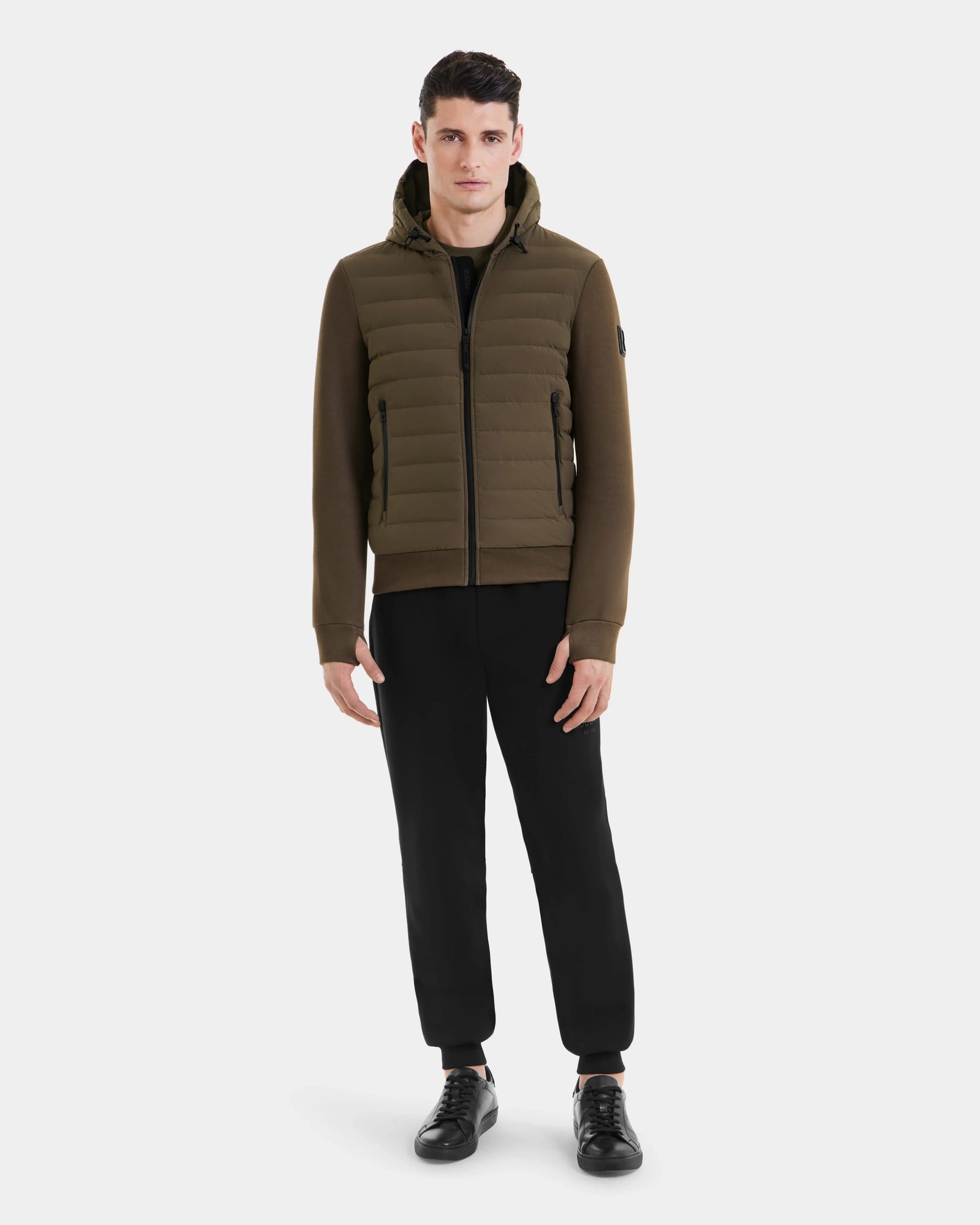A man faces forward against a white background, wearing the Rudsak DEE MAT - 6124028 Army mens khaki green lightweight hybrid down jacket with a hood, black pants, and black sneakers.