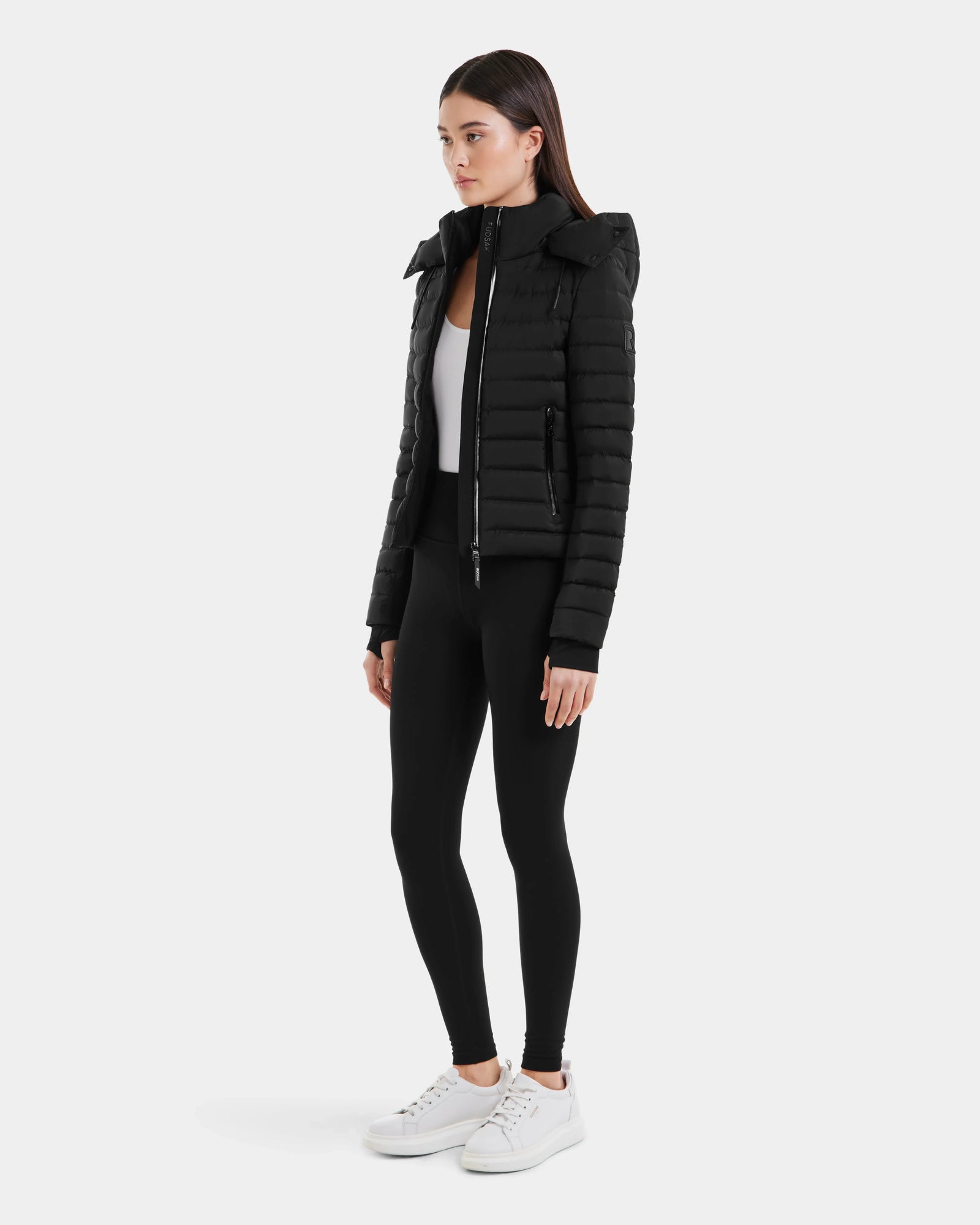 A woman stands slightly to the side against a plain white background, wearing the Rudsak MARYA M - L123559 Black women’s down puffer jacket, black leggings, a white top, and white sneakers.