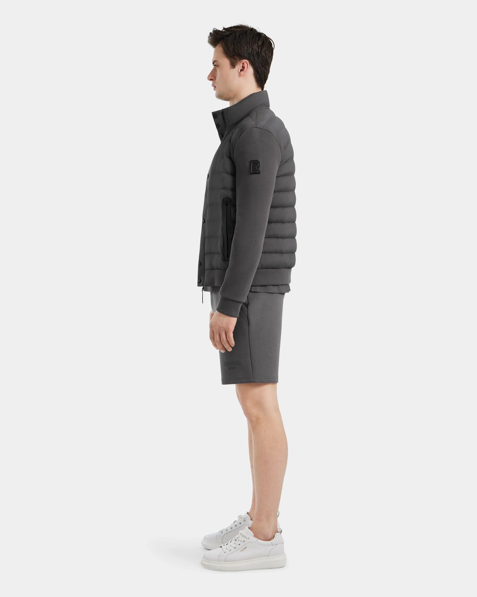 A man stands in profile against a plain background, wearing the Rudsak RAGAB - 6124577 Grey mens hybrid jacket with lightweight down insulation, matching shorts, and white sneakers—ideal for transitional weather.