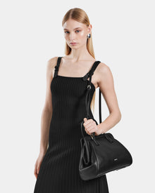 A woman with straight blonde hair wears a sleeveless black dress and silver earrings, holding the Rudsak TAULLA - 8325002 Black pebble leather satchel with removable shoulder strap against a plain white background.