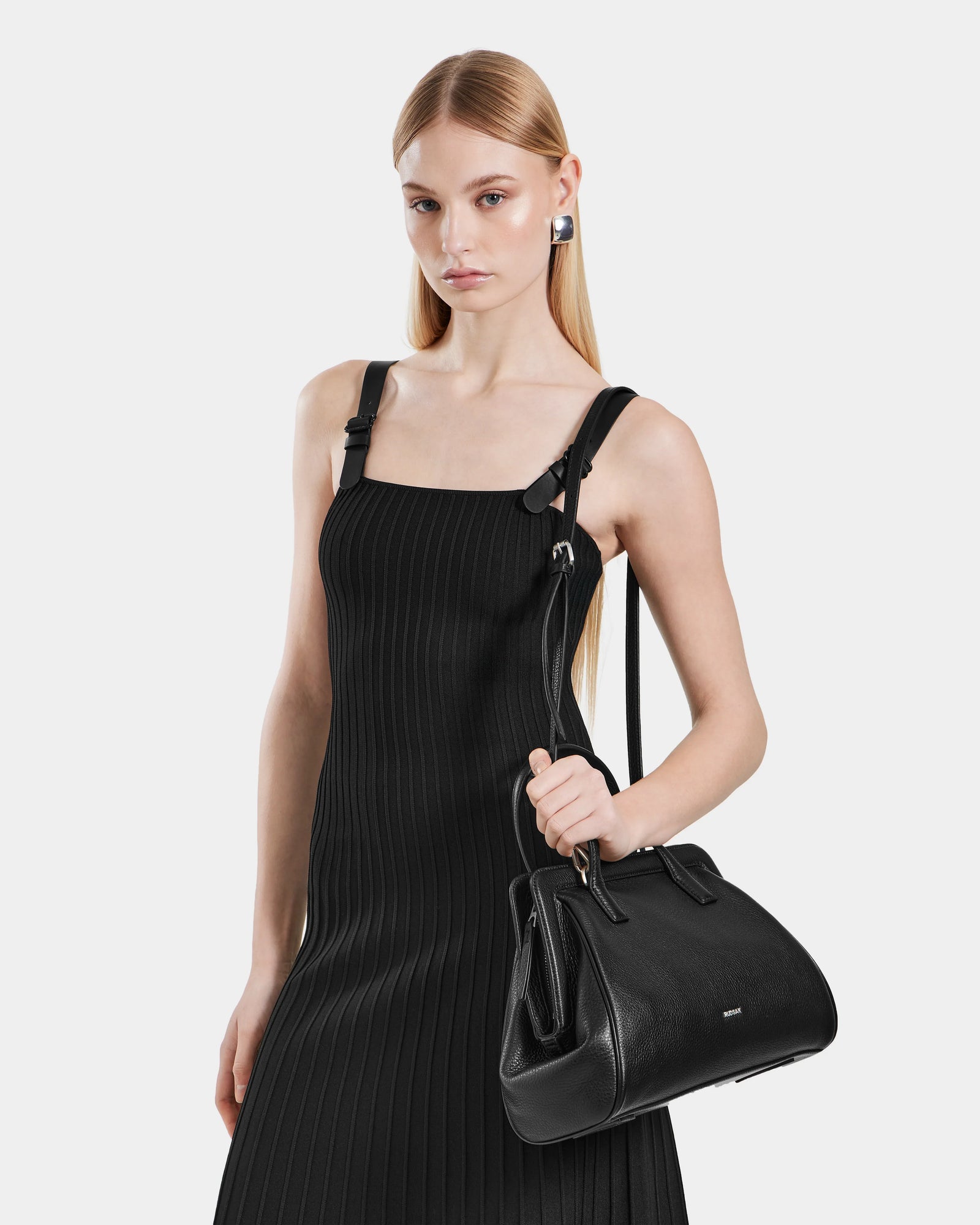 A woman with straight blonde hair wears a sleeveless black dress and silver earrings, holding the Rudsak TAULLA - 8325002 Black pebble leather satchel with removable shoulder strap against a plain white background.