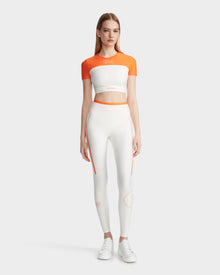 A woman faces forward wearing the Rudsak LILIANE - 8524018 Eggshell women’s fitted crop top and matching high-waisted leggings in white and orange, made from high-performance stretch fabric, paired with white sneakers against a plain light background.