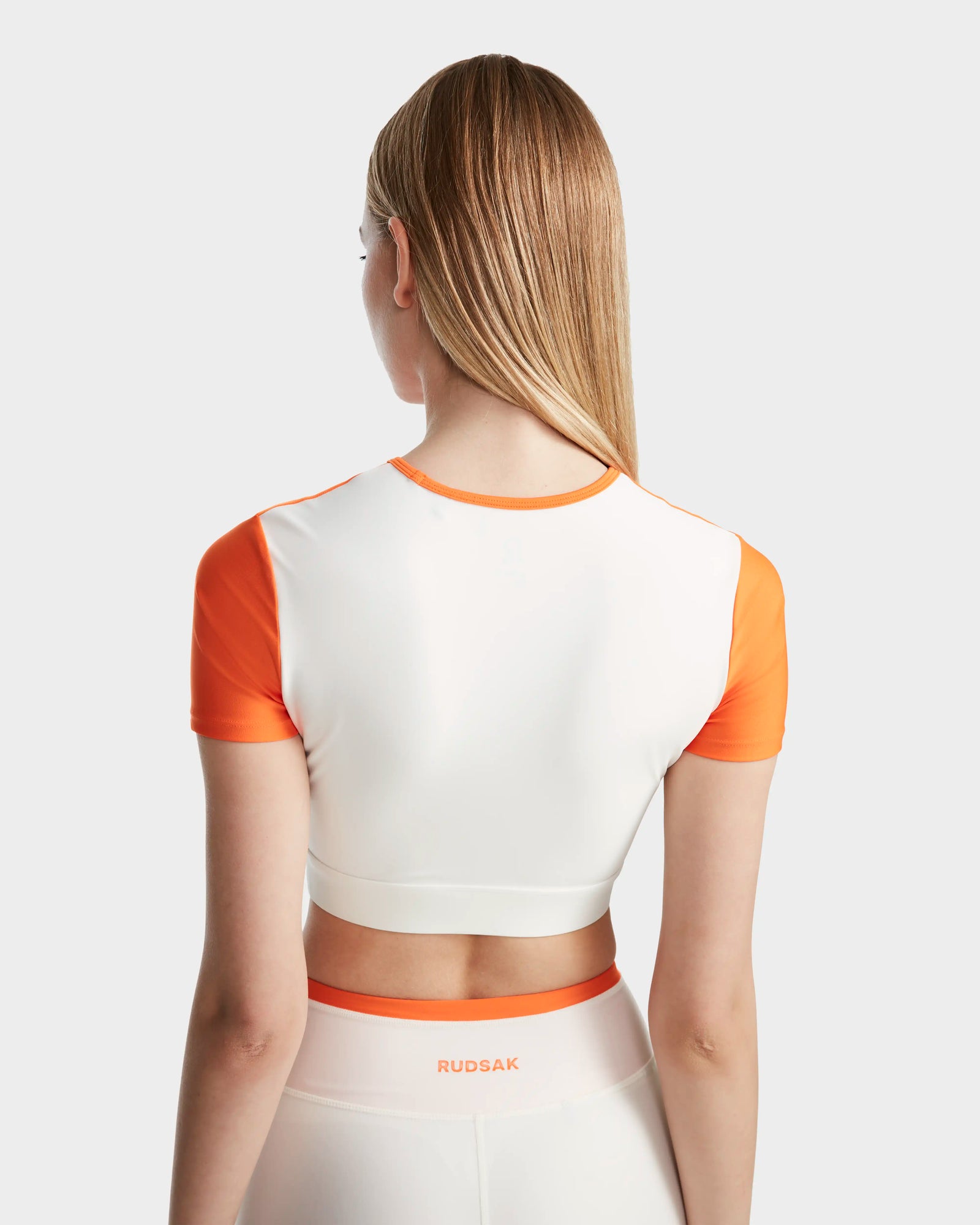A woman with straight, light brown hair is seen from behind wearing the Rudsak LILIANE - 8524018 Eggshell fitted crop top and matching high-waisted leggings in white and orange, featuring stretch fabric and RUDSAK branding on the waistband.