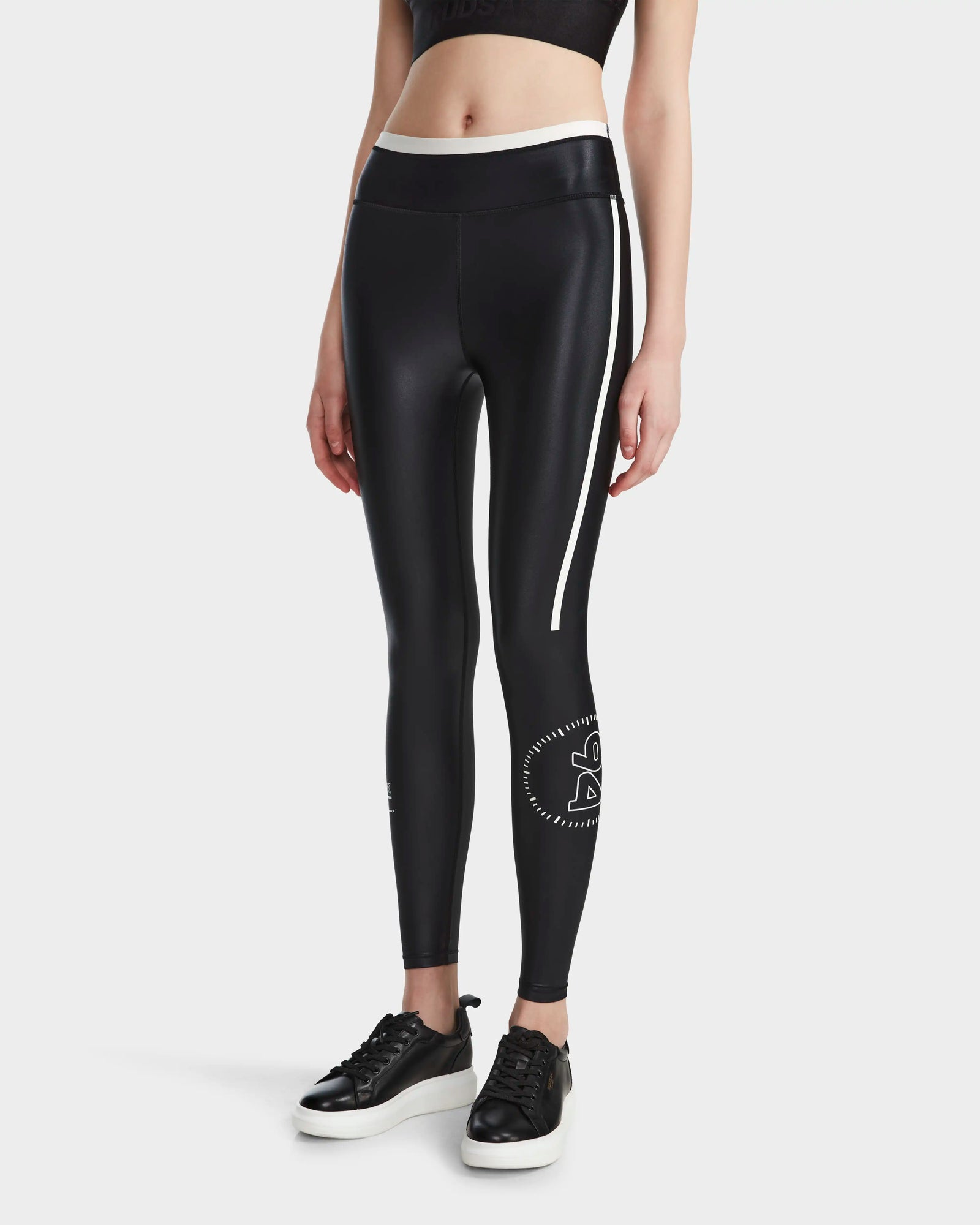 A person wears Rudsaks TAYLA - 8524015 Black high-performance womens leggings with a white stripe and graphic, styled with black sneakers and a black crop top against a plain white background.