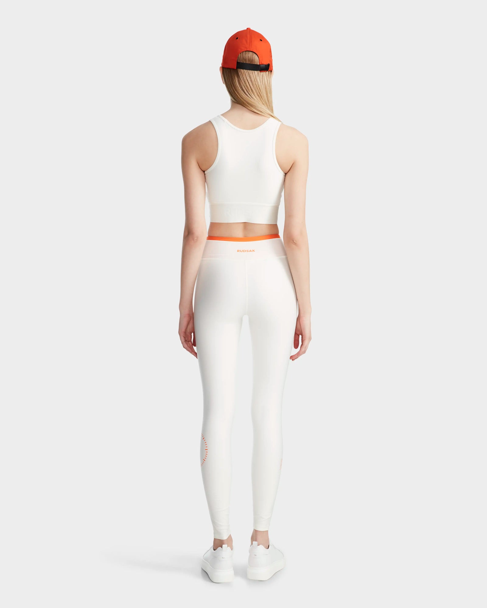 A woman stands facing away, wearing the Rudsak TRACY - 8524016 Eggshell racer-back sports bra and matching leggings, with an orange waistband, white sneakers, an orange cap, and long blonde hair against a light gray background.