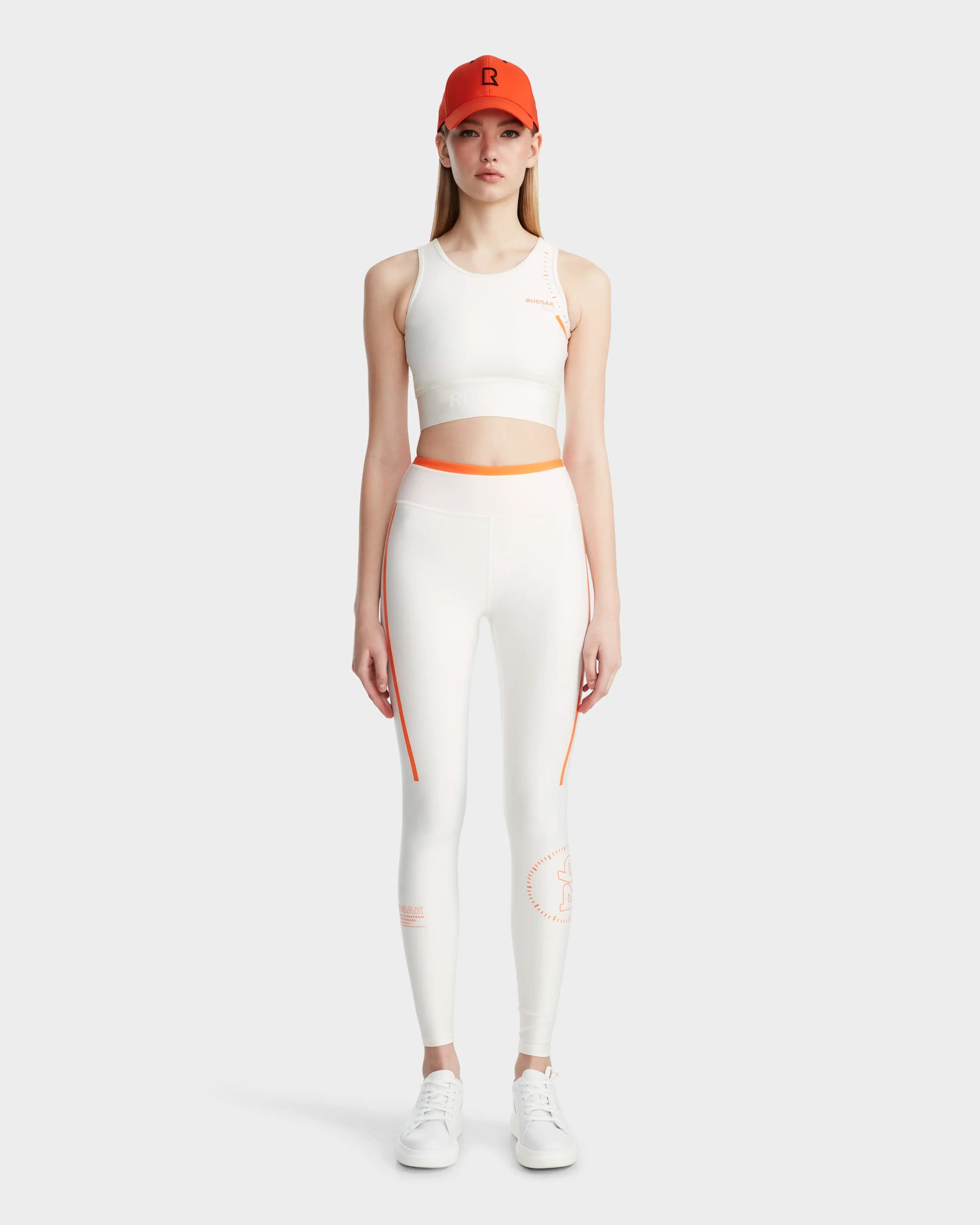 A woman faces forward wearing the Rudsak TRACY - 8524016 Eggshell quick dry sports bra and high-waisted leggings with orange accents, paired with white sneakers and an orange baseball cap. The background is light-colored and plain.