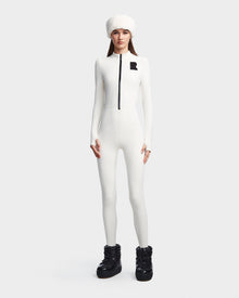 A woman wears the DAVINA - 8523534 Iceland by Rudsak: a fitted white ski suit with front zipper, fleece lining, black R logo on the chest, paired with a white fur hat and black snow boots, standing against a plain background.