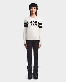 A woman faces forward wearing the ODESSA - 8523540 Iceland soft-knit Rudsak sweater with black sleeve stripes and SKI on the chest, paired with black quilted pants, snow boots, and a furry black hat for winter warmth.