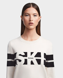A woman with straight brown hair and a neutral expression wears the Rudsak ODESSA - 8523540 Iceland après-ski sweater, featuring soft white knit with black horizontal stripes and RUDSAK printed across the chest, against a light gray background.