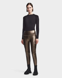 A woman faces forward wearing a fitted black long-sleeve top and Rudsak SIEN P - 8523027 METALLIC CHOCOLATE stretch vegan leather cargo leggings with side pockets, paired with black socks and sneakers, set against a light plain background.