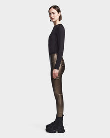 A woman in profile wears a fitted black long-sleeve top, Rudsak’s SIEN P - 8523027 METALLIC CHOCOLATE stretch vegan leather leggings, and black chunky sneakers against a plain light background.