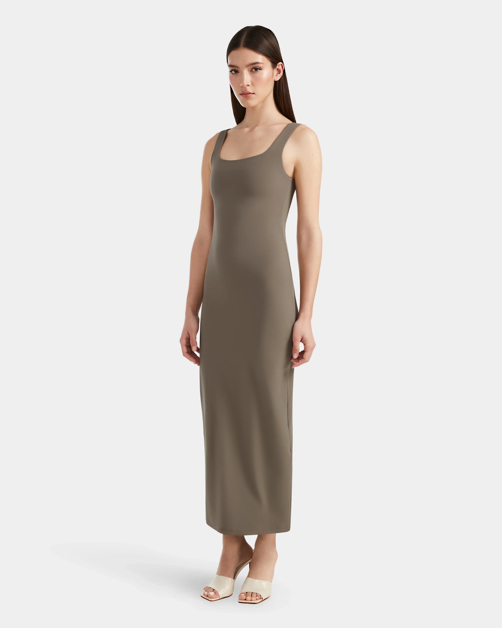 A woman with straight brown hair wears the Rudsak WANDA - 8524035 Cement women’s maxi dress and light open-toe heels, standing against a plain white background, radiating contemporary elegance.