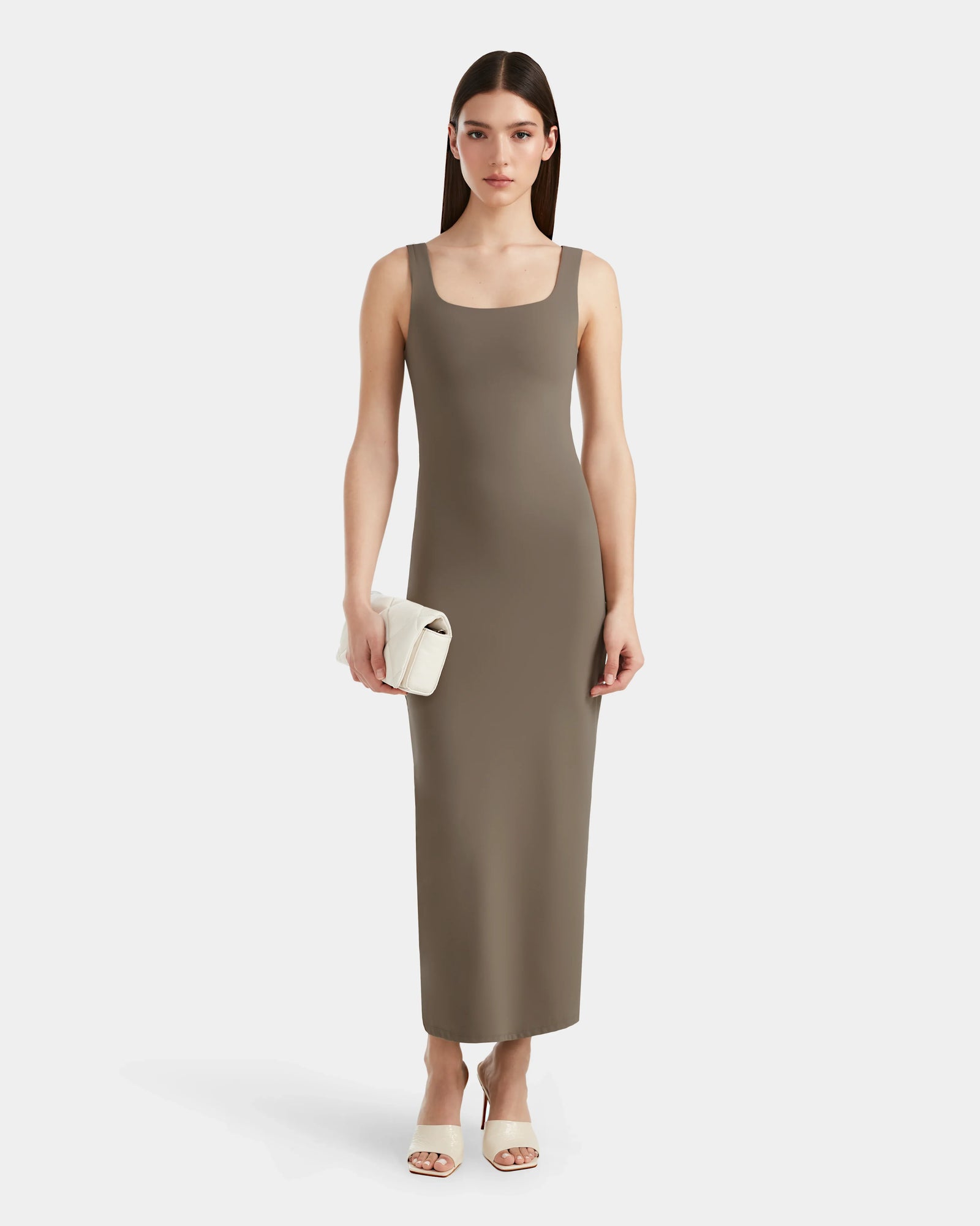 A woman faces forward, exuding modern elegance in the Rudsak WANDA - 8524035 Cement maxi dress, crafted from stretch performance fabric. She holds a white clutch and wears white heeled sandals against a plain white background.