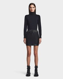 A woman with straight brown hair and a neutral expression faces forward in a black turtleneck, RUDSAK’s PORTIA - 8523033 BLACK vegan leather mini skirt, and black ankle boots, standing against a plain white background.
