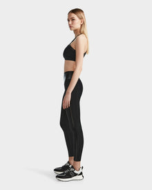 A woman stands in profile wearing the RUDSAK ARYA - 8523002 BLACK sports bra, paired with high-waisted black leggings and black sneakers with white soles, set against a plain white background.