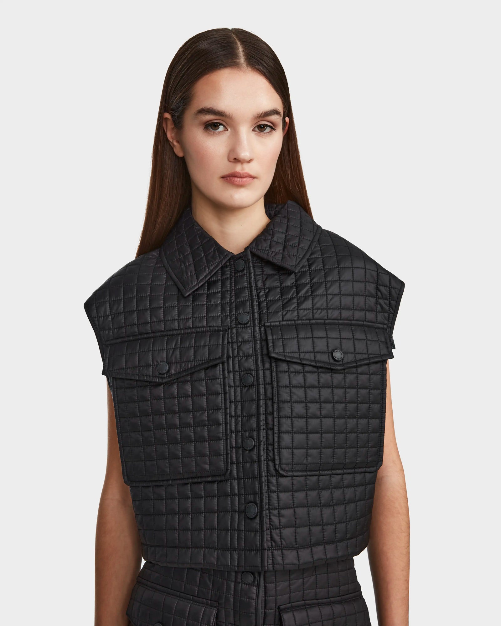 A woman with long brown hair wears the Rudsak SOLSA - 8124048 womens sleeveless black cropped quilted vest with a collar and front pockets, standing against a plain white background.