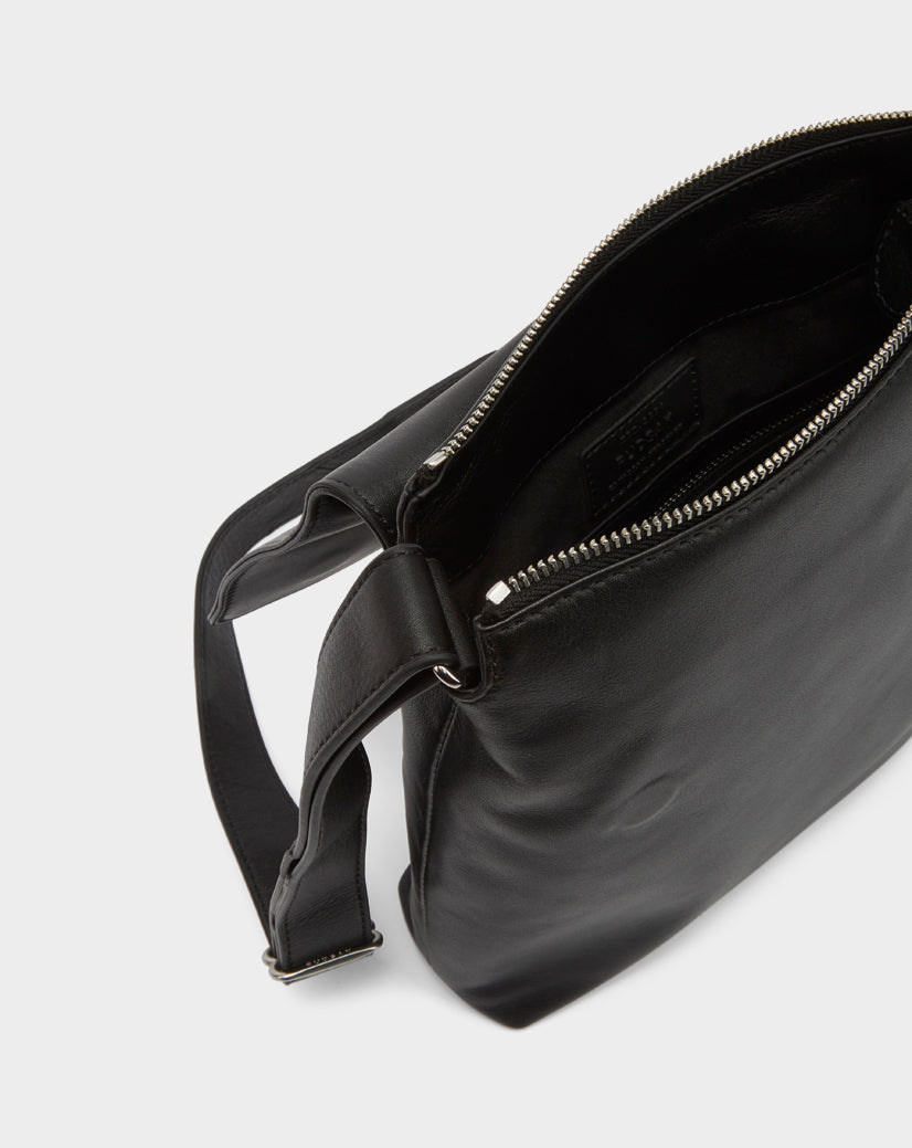 The Rudsak MADDY - 8322512 BLACK crossbody bag features black leather, a silver zipper, an open main compartment with a dark fabric interior, and an adjustable strap—ideal for minimalist style lovers.