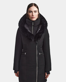 A woman with straight, dark, center-parted hair faces forward wearing the Rudsak CORA - L123538 Black parka. The sustainable coat features a fur-lined hood, zippered pockets, high collar, and is shown against a plain white background.