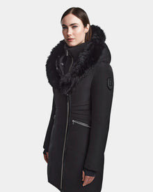 A woman with straight brown hair wears the Rudsak CORA - L123538 Black women's fitted parka, a long black winter coat with a large furry hood and zippered pockets, standing against a plain light background.