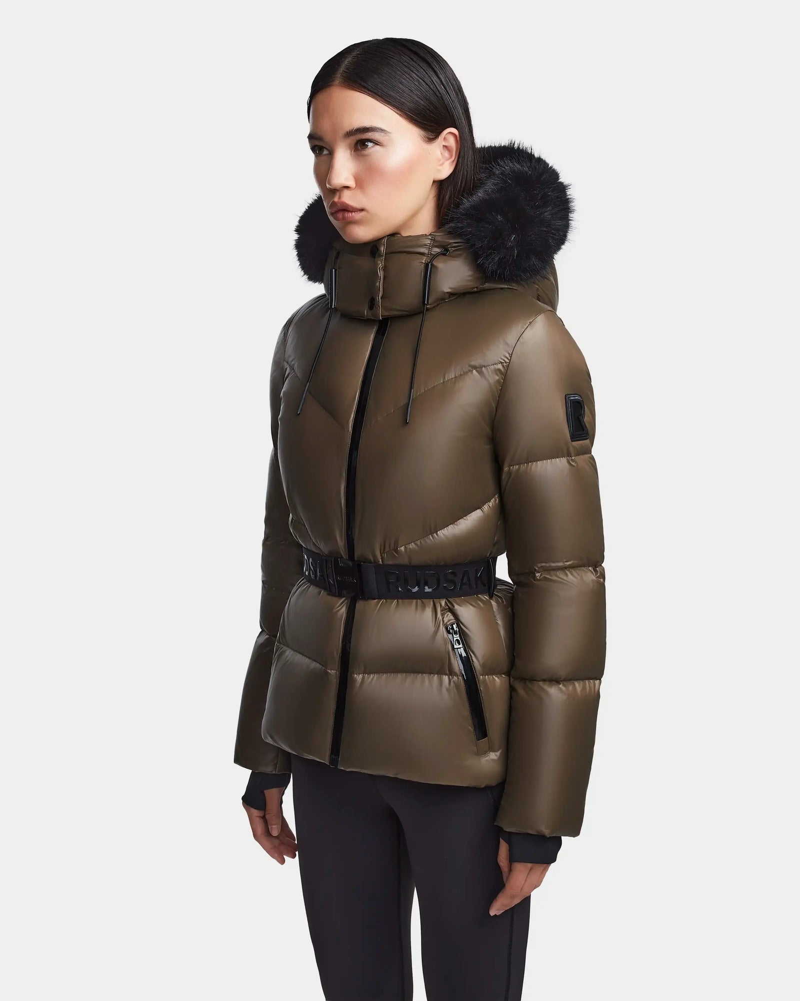 A woman wears the Rudsak CHRISTELLE S - L123524 women's chevron quilted down puffer in light olive, featuring a black fur-trimmed hood, black belt, and zipper details, standing against a plain light gray background.